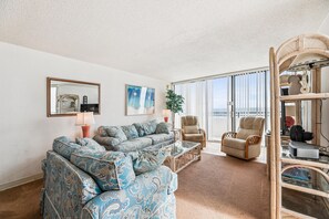 Sala de estar com sofá, poltrona e mesa de centro de vidro. Há uma janela grande com persianas e vista para o exterior.