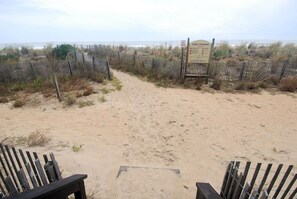 Un camino de arena que lleva a un paso por las dunas, rodeado de cercas de madera y vegetación escasa.