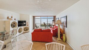 A modern living room with a red sofa, a dining table with chairs, a TV, and a balcony with a view. - Includes Xplorie Activities and Linens. PYRAMID - 2BR oceanfront condo w/outdoor pool. (Ocean City)