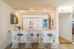 A kitchen with a bar area, white bar stools, and a painting of a beach scene. - Includes Xplorie Activities and Linens. PYRAMID - 2BR oceanfront condo w/outdoor pool. (Ocean City)