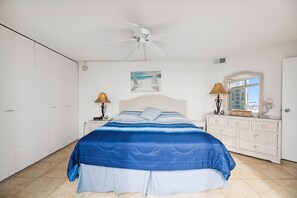 Een slaapkamer met een bed, een plafondventilator, een schilderij, een kaptafel en een raam.