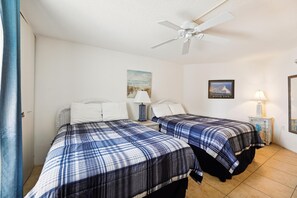 A hotel room with two beds, a ceiling fan, a painting of a beach, and a framed picture of a coastal city.
