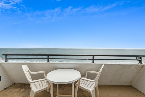 A balcony with a white table and two chairs, overlooking a beach with waves.