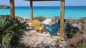 Un quiosco de madera con dos sillas, una amarilla y una azul, da vista a una playa con aguas turquesas.