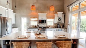 Una cocina amplia con una isla grande, armarios blancos y tres taburetes de mimbre.