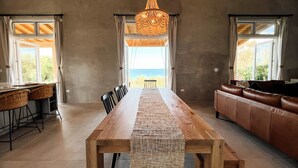Una habitación amplia con una larga mesa de madera, un sofá de cuero y vistas al mar a través de grandes ventanales.
