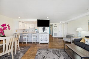 Un salon moderne avec un canapé, un coin repas avec des chaises, une cuisine avec des meubles blancs et un téléviseur à écran plat fixé au mur.