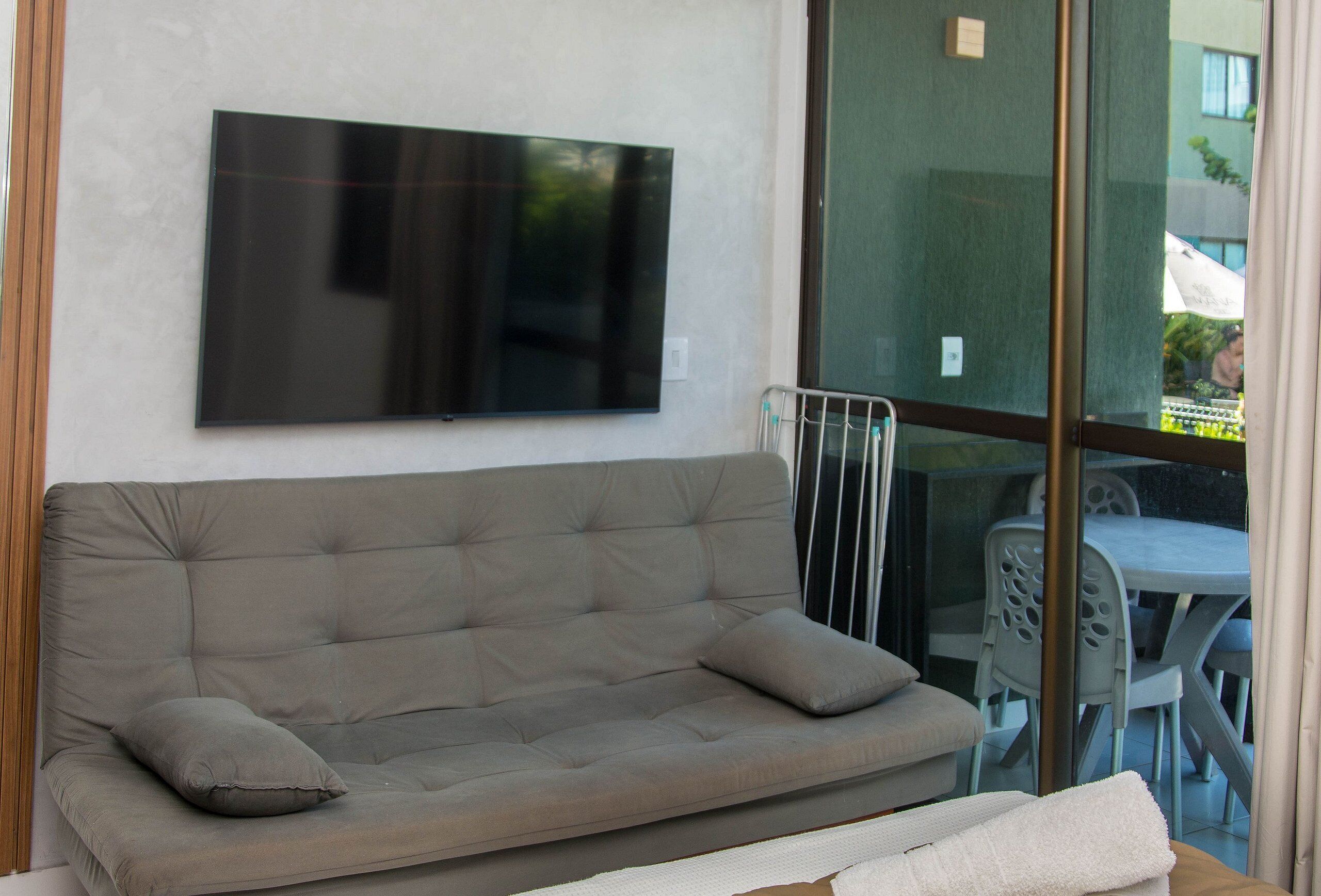 A grey sofa with two cushions, a flat-screen TV mounted on the wall, and a glass door leading to an outdoor area with a table and chairs.
