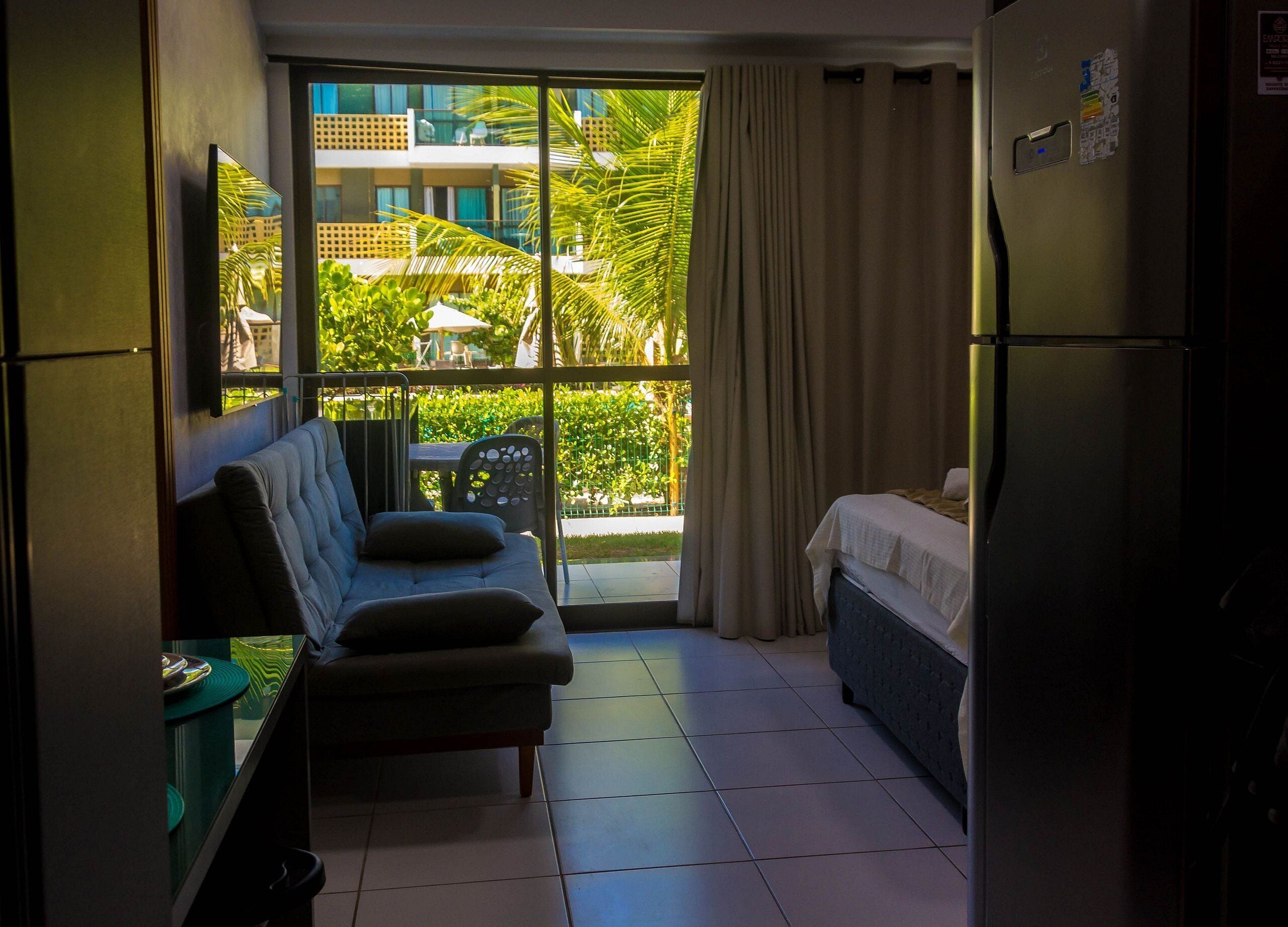 A compact hotel room with a balcony, a sofa, a bed, and a refrigerator.