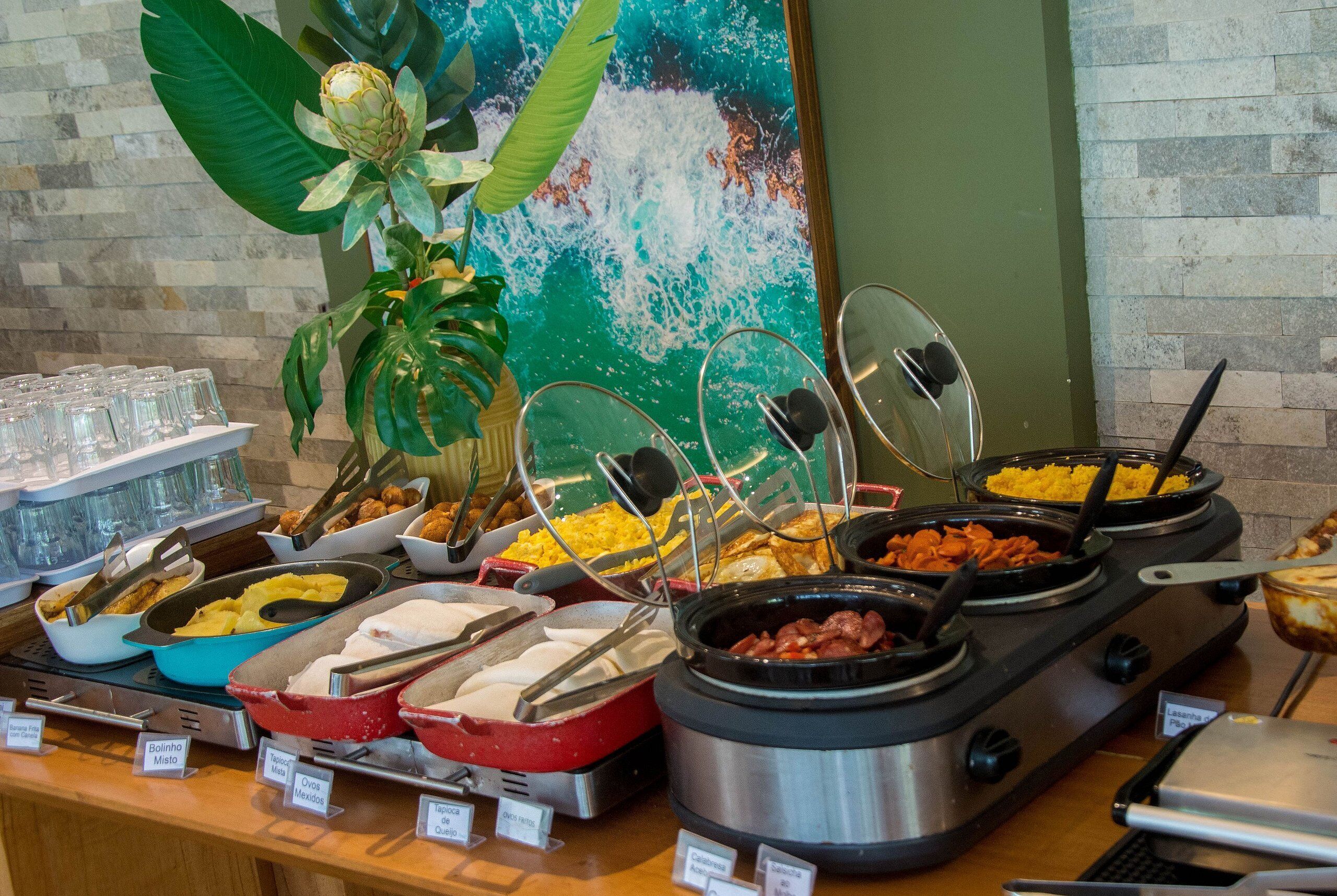 A buffet with various food options, including a red dish labeled "Bolinho Misto," a blue dish labeled "Ovo Mexida," and a black dish labeled "Laranja Pica."
