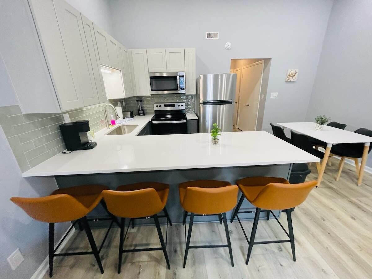 A modern kitchen with white cabinets, a stainless steel refrigerator, a microwave, and a stove. There are three orange bar stools and a white island.