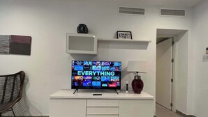 Una sala de estar moderna con un televisor de pantalla plana mostrando la interfaz de un servicio de streaming, un mueble de entretenimiento blanco con una lámpara y un cuadro en un estante.
