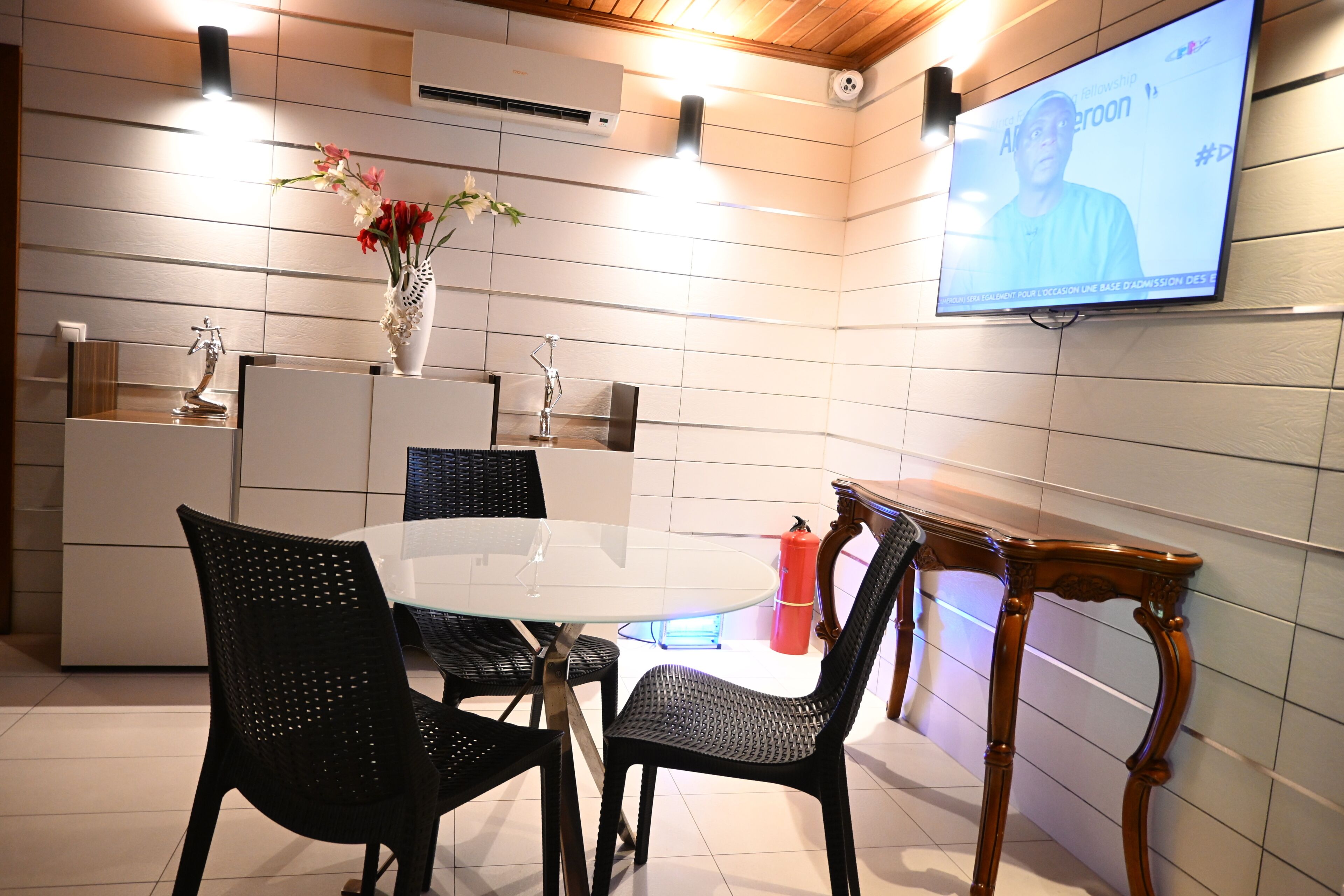 Un comedor moderno con una mesa redonda, sillas negras, un florero con flores y una pantalla de televisión que muestra a una persona.