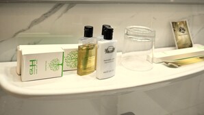 Una encimera de baño con artículos de aseo, incluyendo una caja con la etiqueta 'Grand Lux Hotel', una botella de 'Gel de ducha Verbena Limón', una botella de 'Loción corporal Verbena Limón', un vaso de agua y una tarjeta.