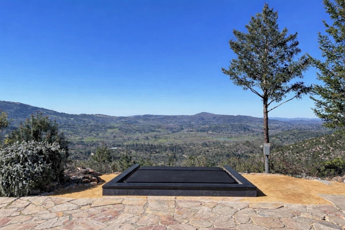 A stone patio with a black rectangular platform, surrounded by natural vegetation and a clear view of distant hills.