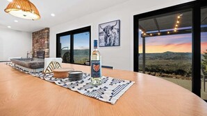 A modern dining area with a wooden table, a bottle of Kenwood whisky, and a view of the landscape through large windows. - When in Glen | Panoramic View | Pool / Hot Tub (Glen Ellen)