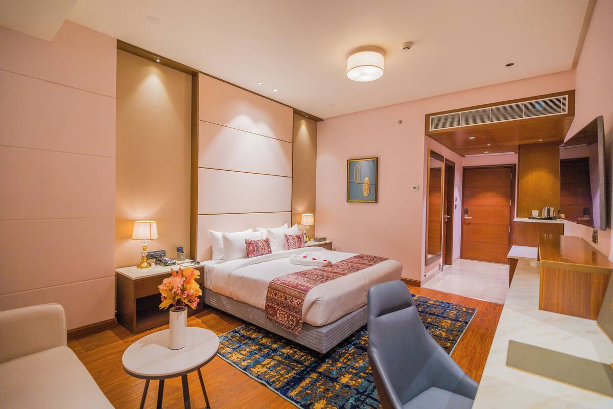 Ein Hotelzimmer mit einem Bett, Nachttischen, einem Schreibtisch, einem Sessel, einem kleinen Tisch mit einer Vase und einem gemusterten Teppich.