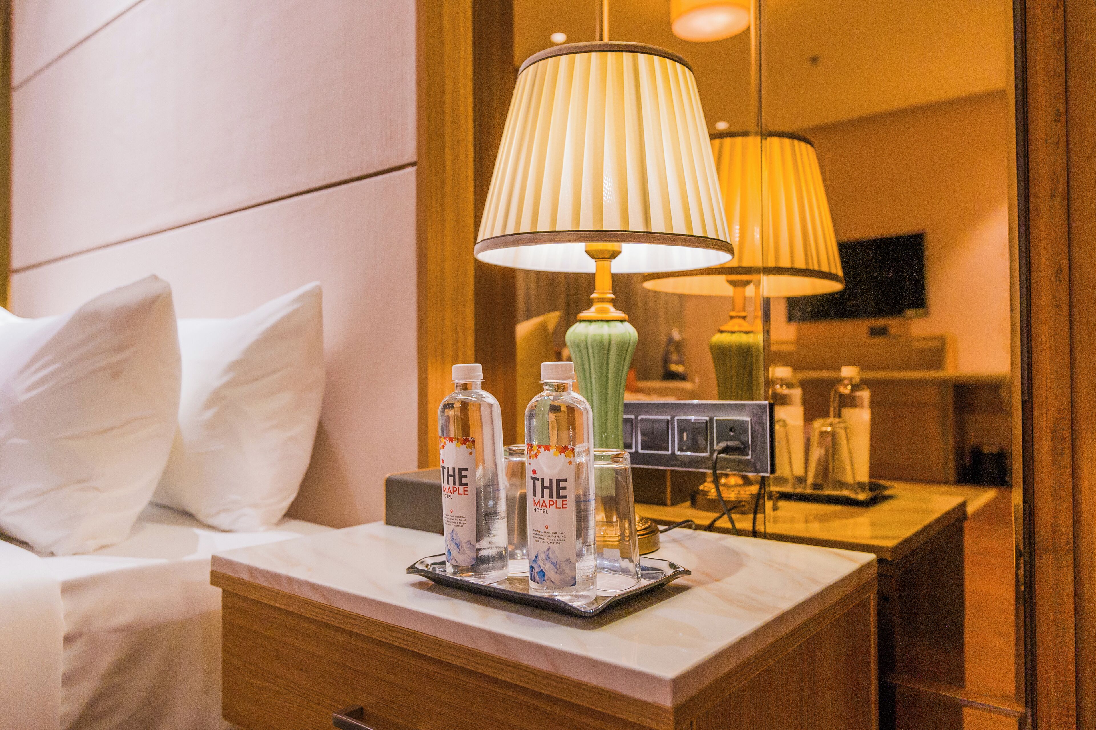 A hotel room with a bed, a nightstand with a lamp, and a tray with water bottles.