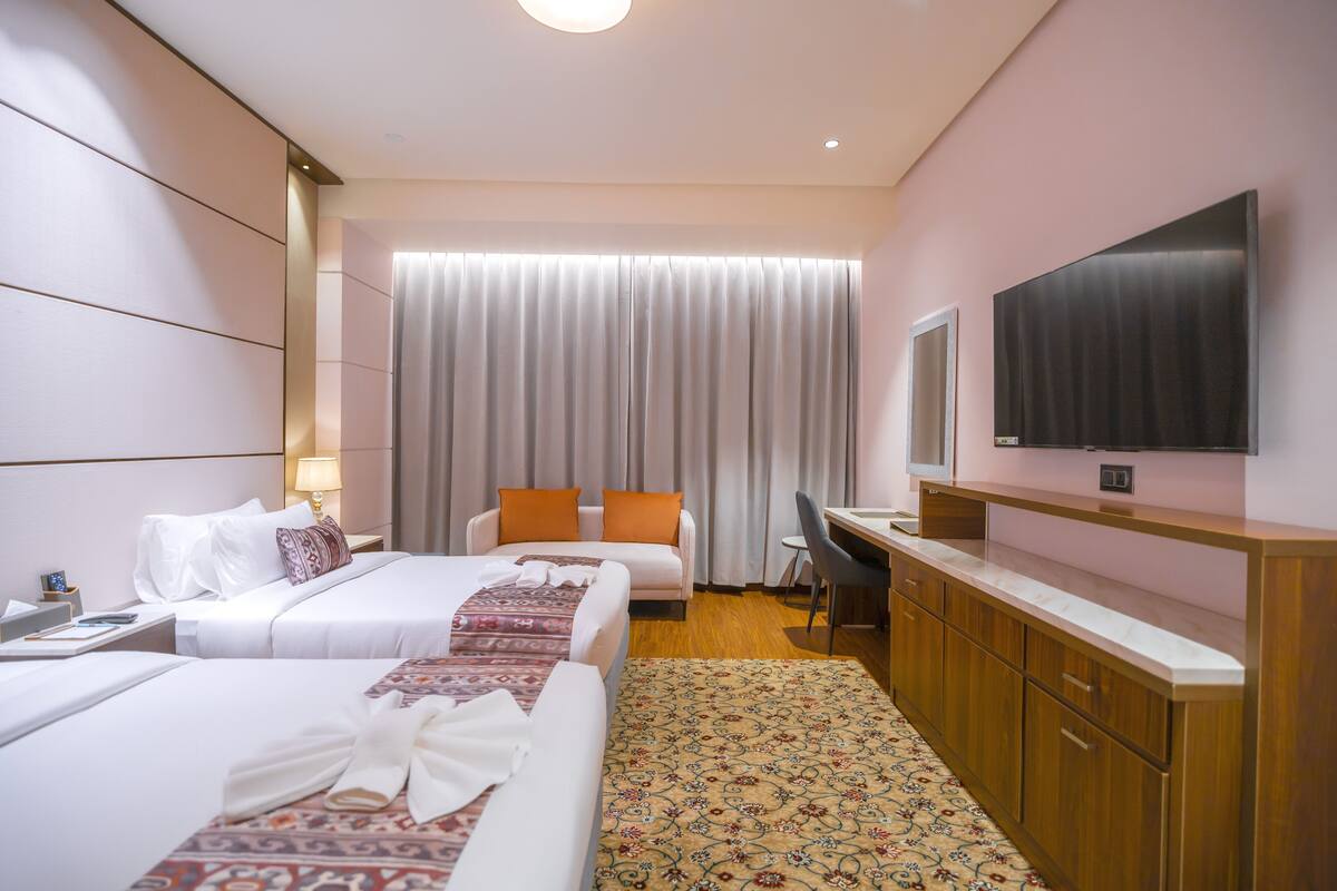 Ein Hotelzimmer mit zwei Betten, einem Schreibtisch, einem Stuhl, einem Flachbildfernseher und einem großen Fenster mit Vorhängen.