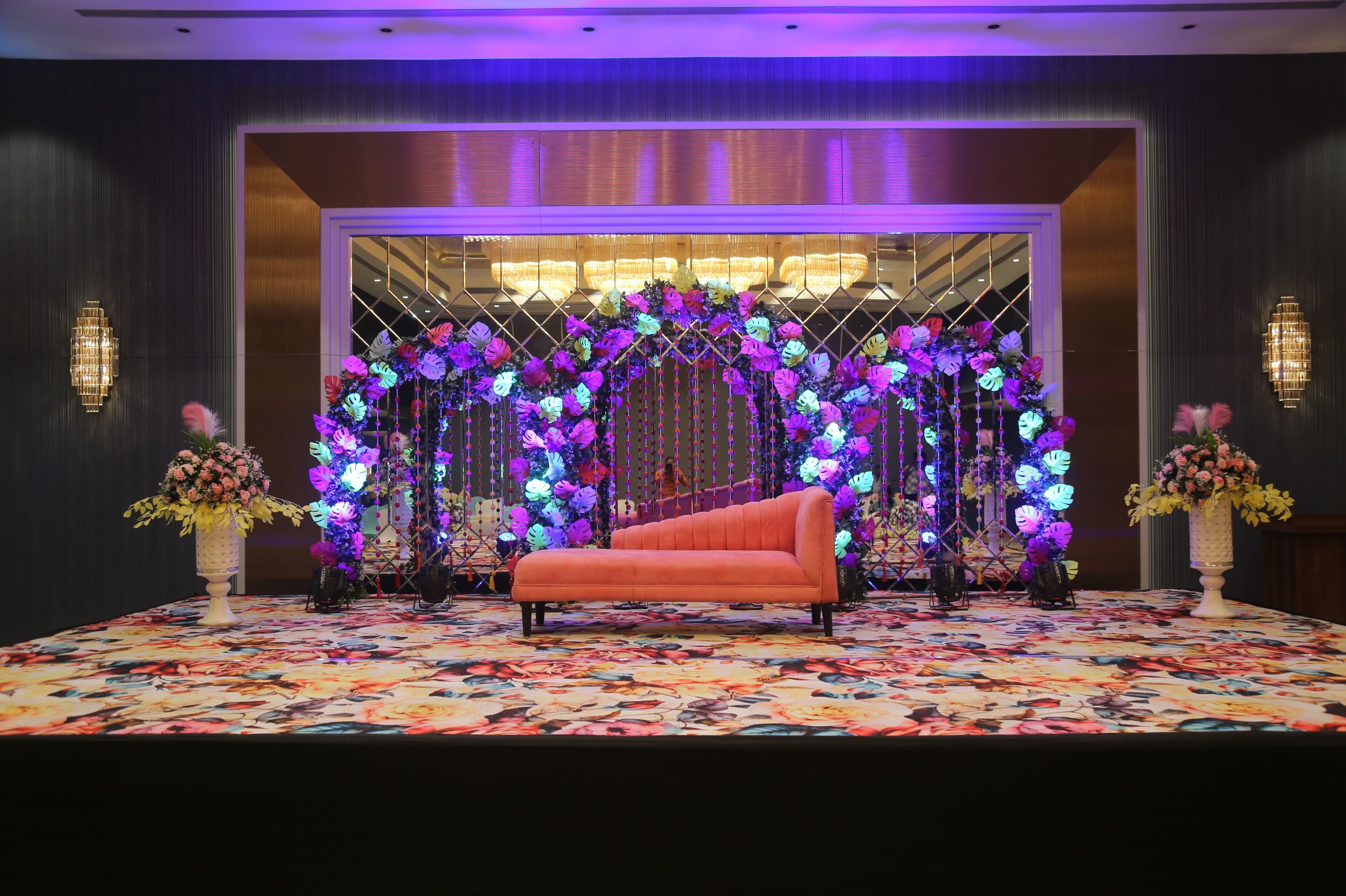 A stage with a floral arch and a sofa, surrounded by wall sconces and vases.