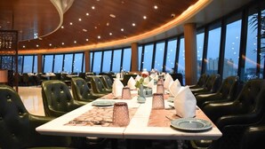 A restaurant with a long table set for a meal, surrounded by chairs, with a city view through large windows.