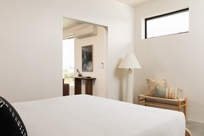 A bedroom with a bed, a bedside table with a lamp, a small wooden bench with cushions, a painting on the wall, and an air conditioning unit.
