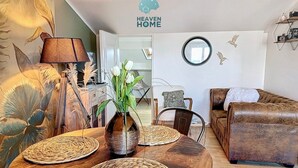 A living room with a wooden table, woven placemats, a vase with flowers, a brown leather sofa, a mirror, and a wall decal.