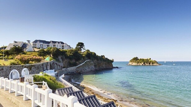 A coastal hotel with a stone wall, a beach with sun loungers, and a view of the sea and a small island.