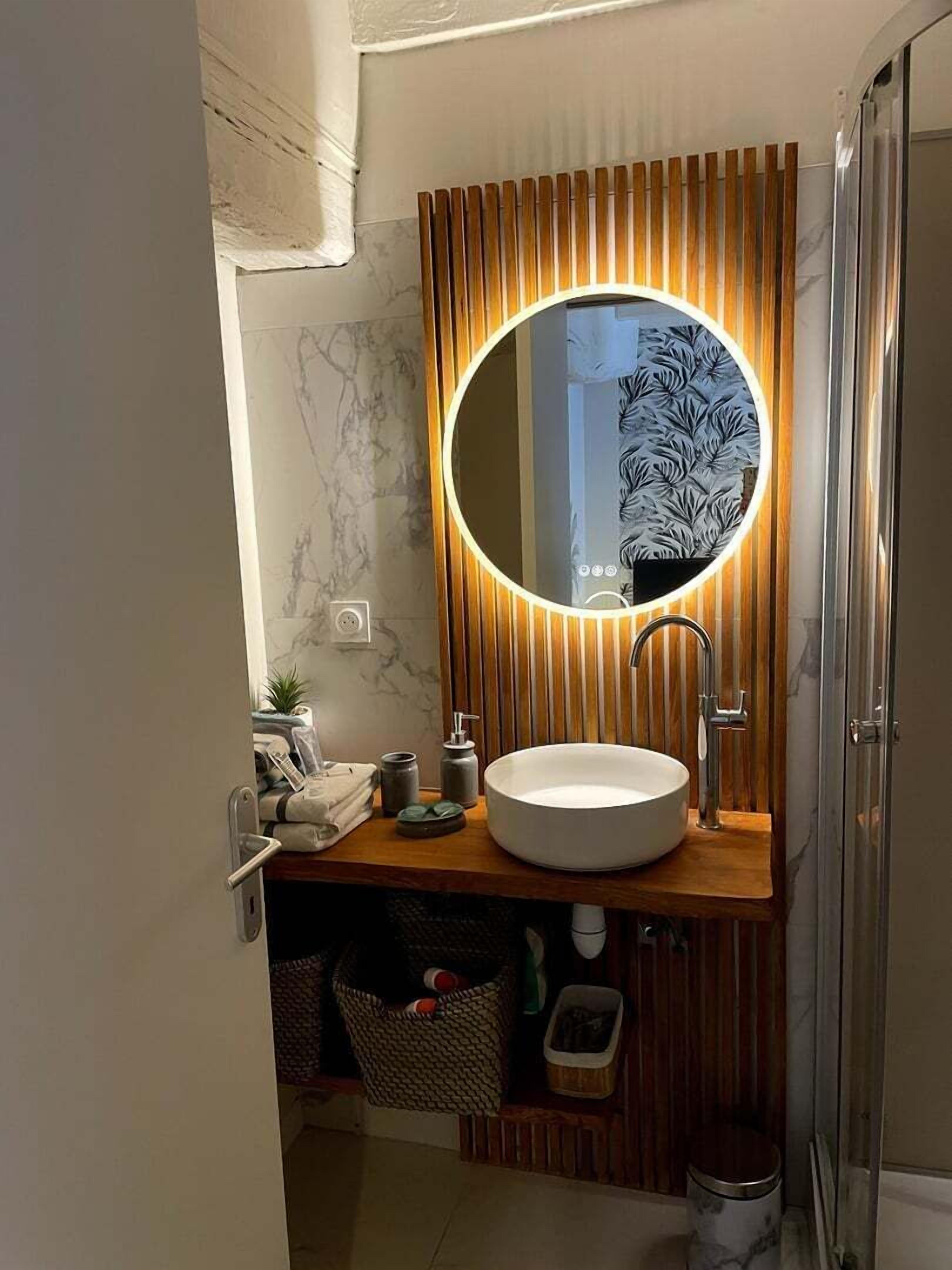 Un baño moderno con tocador de madera, un espejo redondo iluminado, un lavamanos blanco y una zona de ducha.