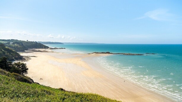 Une plage côtière avec un rivage sablonneux et une mer calme, entourée de verdure et de collines.
