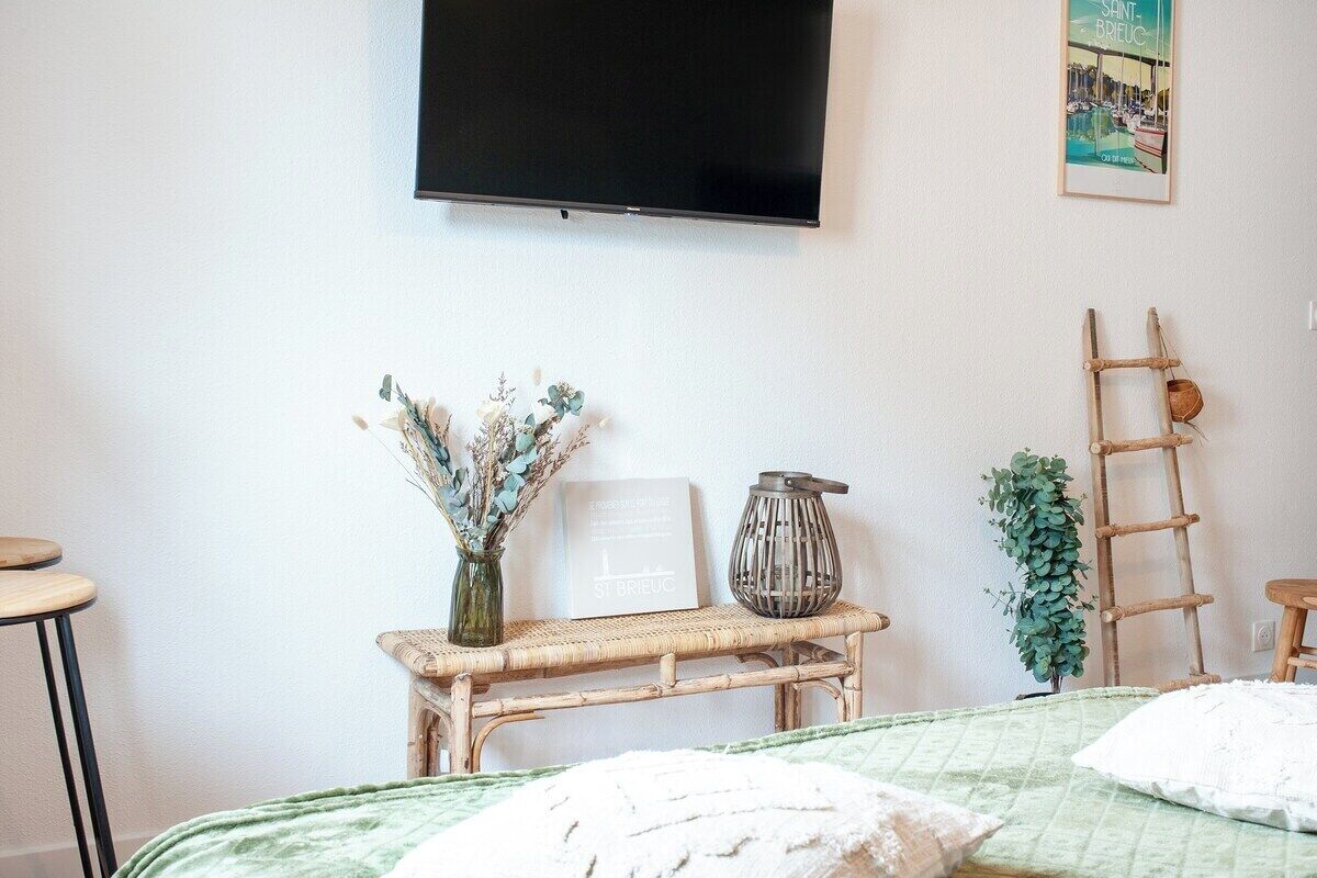 A bedroom with a bed, a television mounted on the wall, a small table with a vase of flowers, a ladder, and a framed picture.