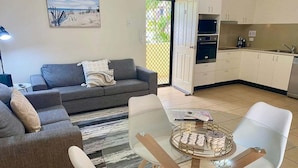 Ein modernes Wohnzimmer mit einer grauen Couch, einem Glastisch und einem Essbereich mit weißen Stühlen. Im Hintergrund ist eine Küche mit weißen Schränken und einem Spülbecken zu sehen.