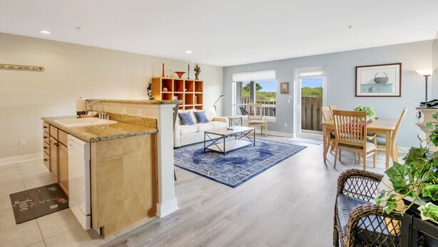 Une cuisine moderne dotée d’un plan de travail en granit, de meubles en bois et d’un lave-vaisselle encastré. On y trouve un coin repas avec une table et des chaises, un tapis bleu et un fauteuil en osier.