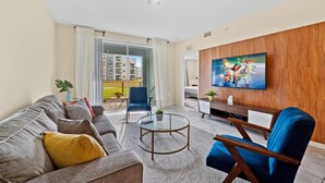 A modern living room with a sofa, armchair, and a coffee table. A flat-screen TV is mounted on the wall, and there is a balcony with a view of buildings. - Minutes From Theme Parks! Free Water Park access! (Kissimmee)