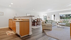 A modern kitchen with wooden cabinets, a built-in dishwasher, and a dining area with a table and chairs. There is a living room with a sofa and a coffee table. - 1313 - Paws in the sand, head in the sky (Westport)