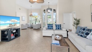 A spacious living room with a large flat-screen TV, a ceiling fan, and a view of the beach through large windows. - The Seahorse House I Wide Views on Tip Lot Minutes from the Gulf (Marco Island)