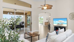 A spacious living area with a large sofa, a coffee table, and a TV displaying a beach scene. There is a ceiling fan, a dining area with chairs, and a view of the outdoors through glass doors. - The Seahorse House I Wide Views on Tip Lot Minutes from the Gulf (Marco Island)