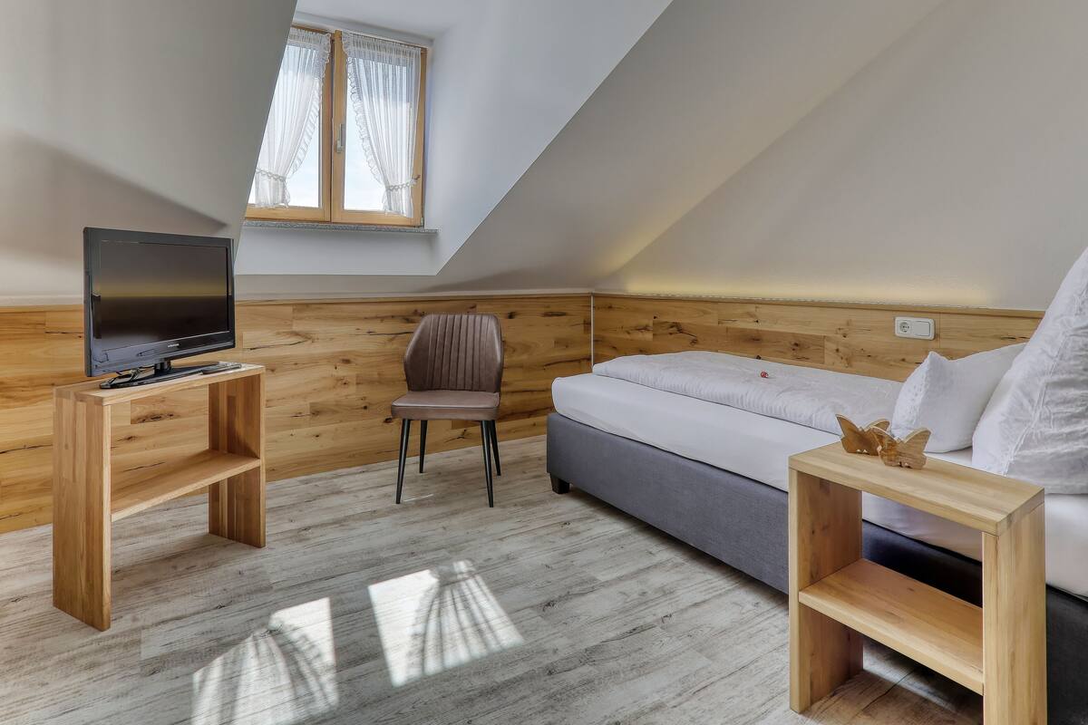 A modern, well-lit room with a wooden floor, a bed, a TV on a wooden stand, and a chair.
