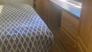 A compact RV interior with a bed, a desk with a computer, wooden cabinets, and a window with a view of greenery. - Cozy motorhome with loads of outdoor activities, farm animals and trails (Stockbridge)