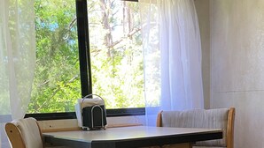 A wooden table with two chairs, a small black object on the table, and a window with a view of trees. - Cozy motorhome with loads of outdoor activities, farm animals and trails (Stockbridge)