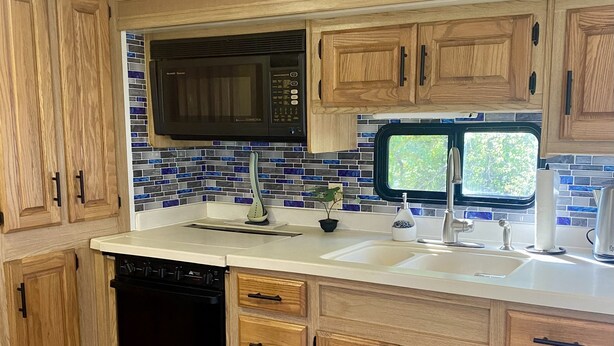 A kitchen with wooden cabinets, a black microwave, a sink with a faucet, and a window. - Cozy motorhome with loads of outdoor activities, farm animals and trails (Stockbridge)