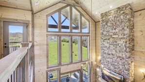 Ein Zimmer mit hohen Decken, großen Fenstern, einem steinernen Kamin und Blick auf eine grüne Landschaft.