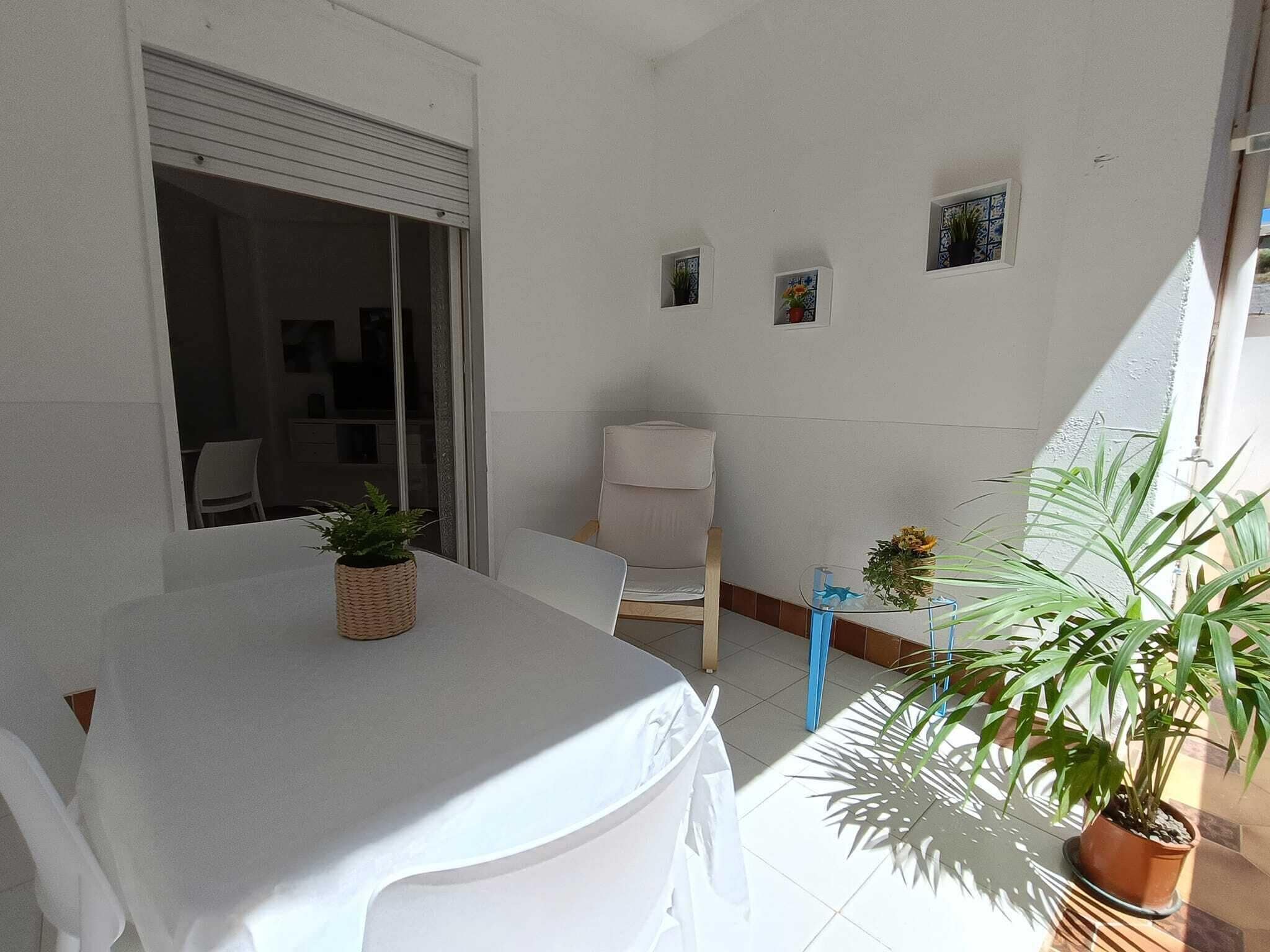 Ein kleines, sauberes Zimmer mit einem weißen Tisch und Stühlen, einer kleinen Topfpflanze und einem Fenster mit Blick.