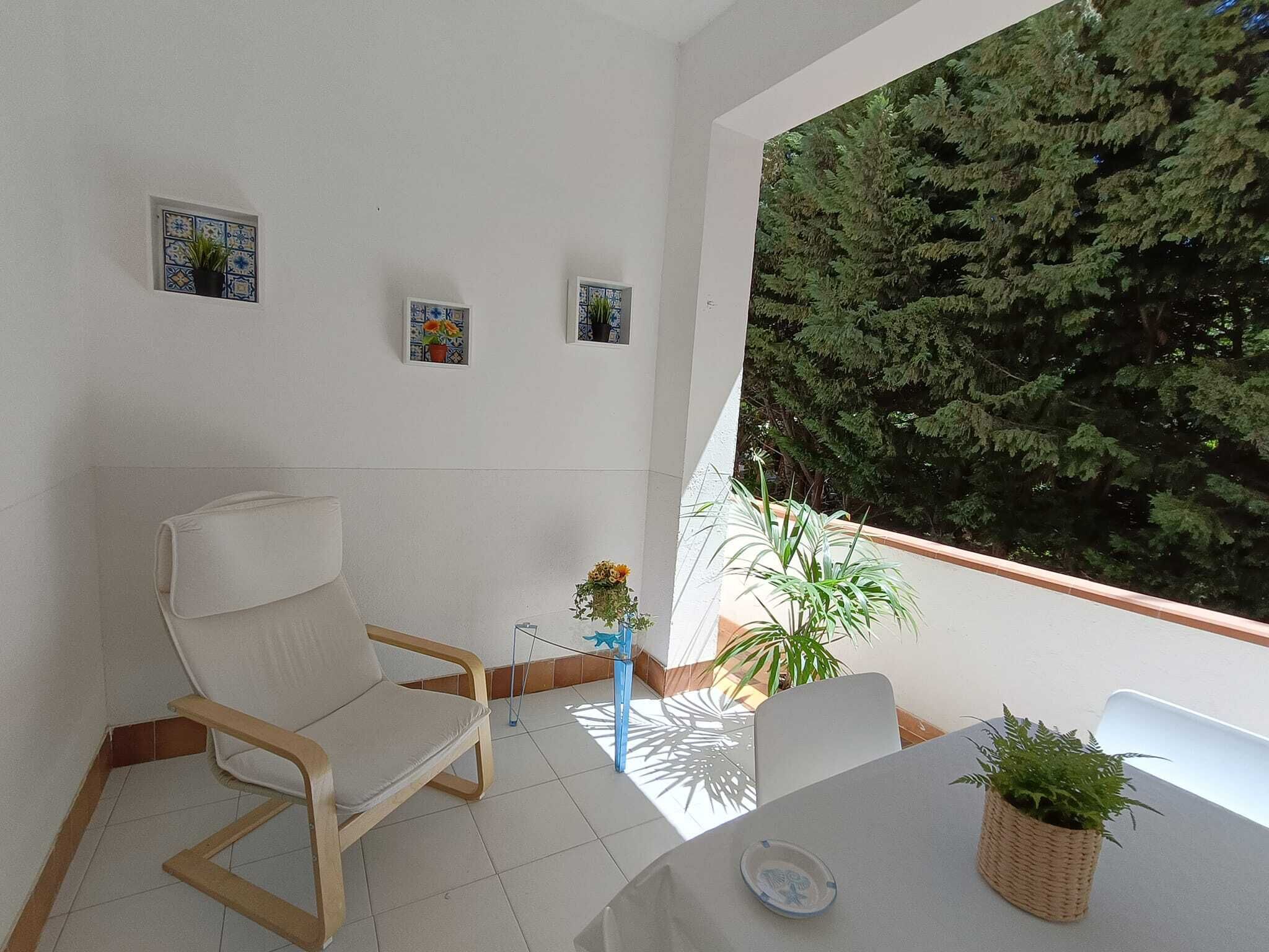 A room with a white chair, a small table, and a view of a dense green tree through a window.