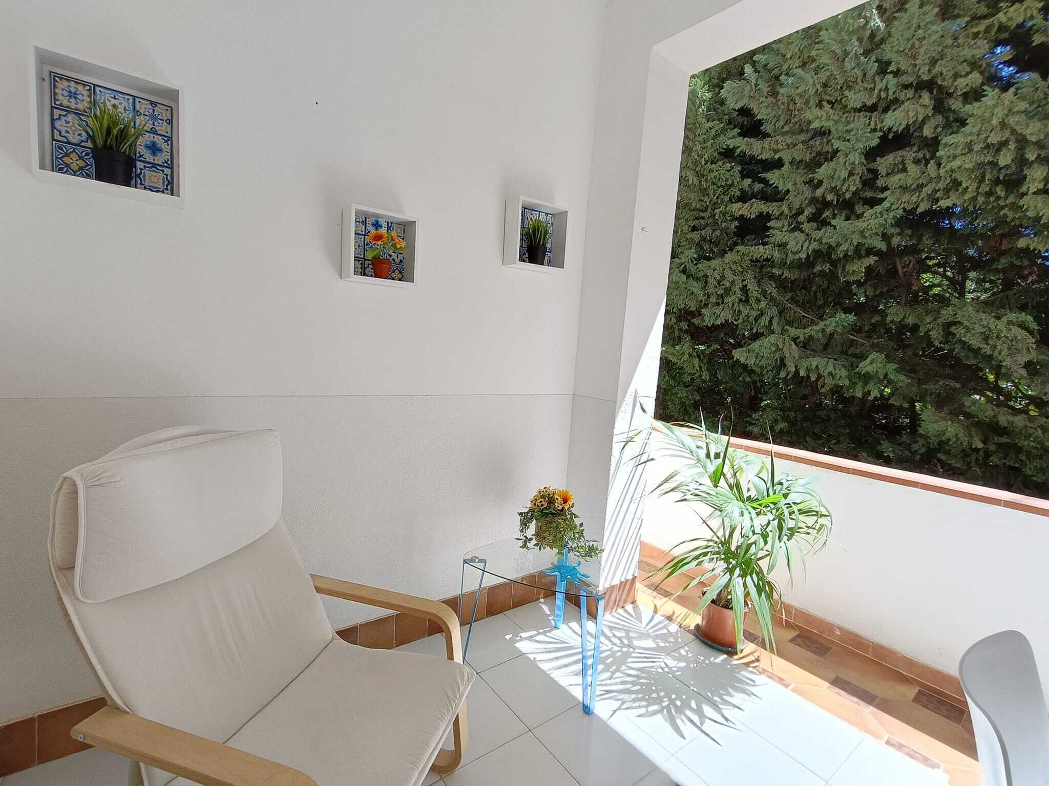 A modern living room with a white sofa, a small table, and a balcony with potted plants and a view of trees.