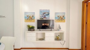 Un salon avec un meuble de rangement blanc pour l’équipement audiovisuel, un téléviseur affichant le logo « NETFLIX », et trois tableaux sur le thème de la plage accrochés au-dessus.