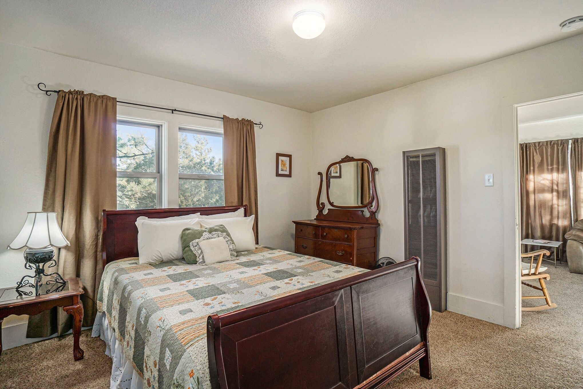 A bedroom with a wooden bed, a nightstand with a lamp, a dresser, a mirror, and a window with curtains.