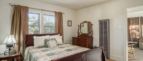A bedroom with a wooden bed, a nightstand with a lamp, a dresser, a mirror, and a window with curtains.