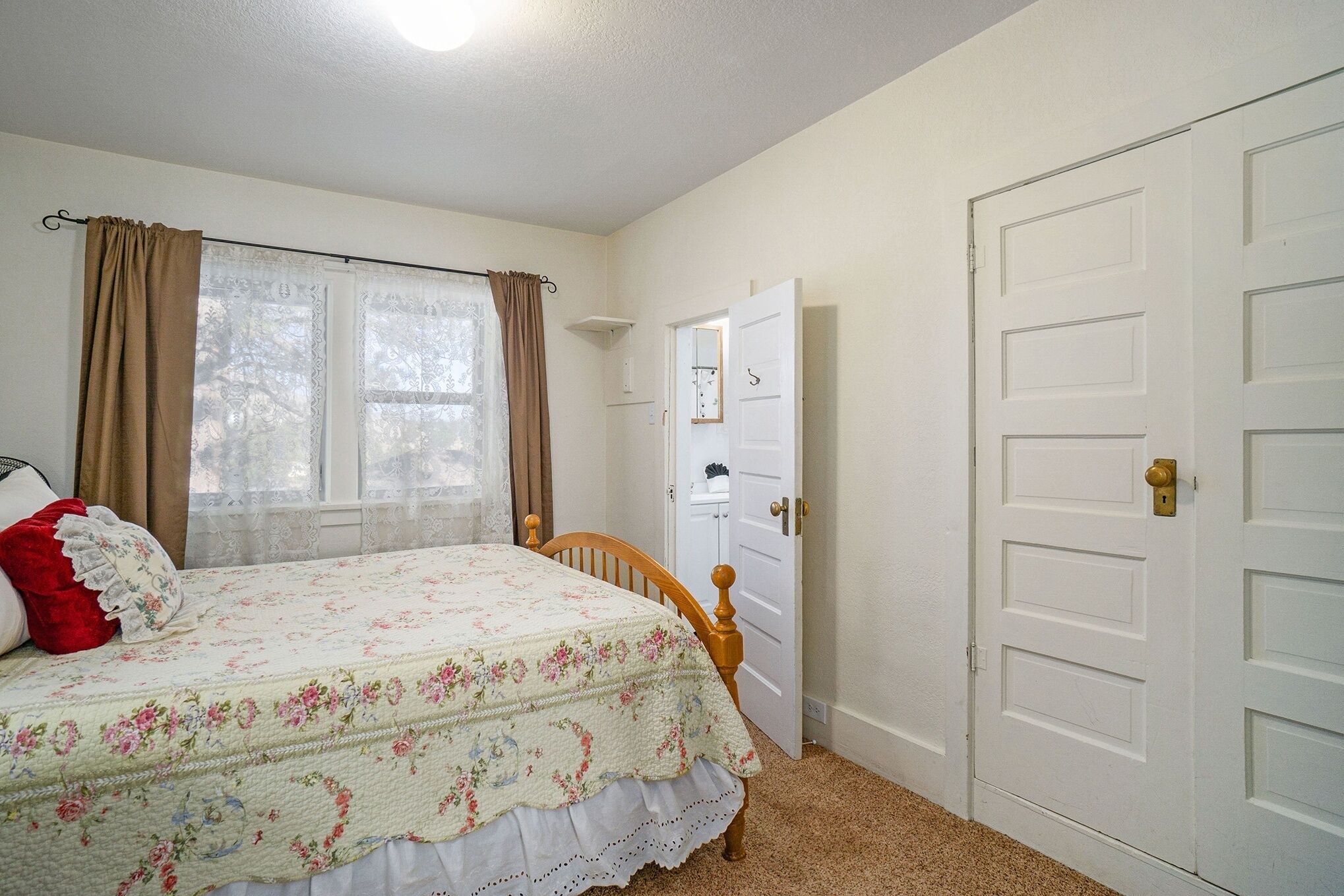 A bedroom with a bed, a window with curtains, a door, and a wall-mounted shelf.