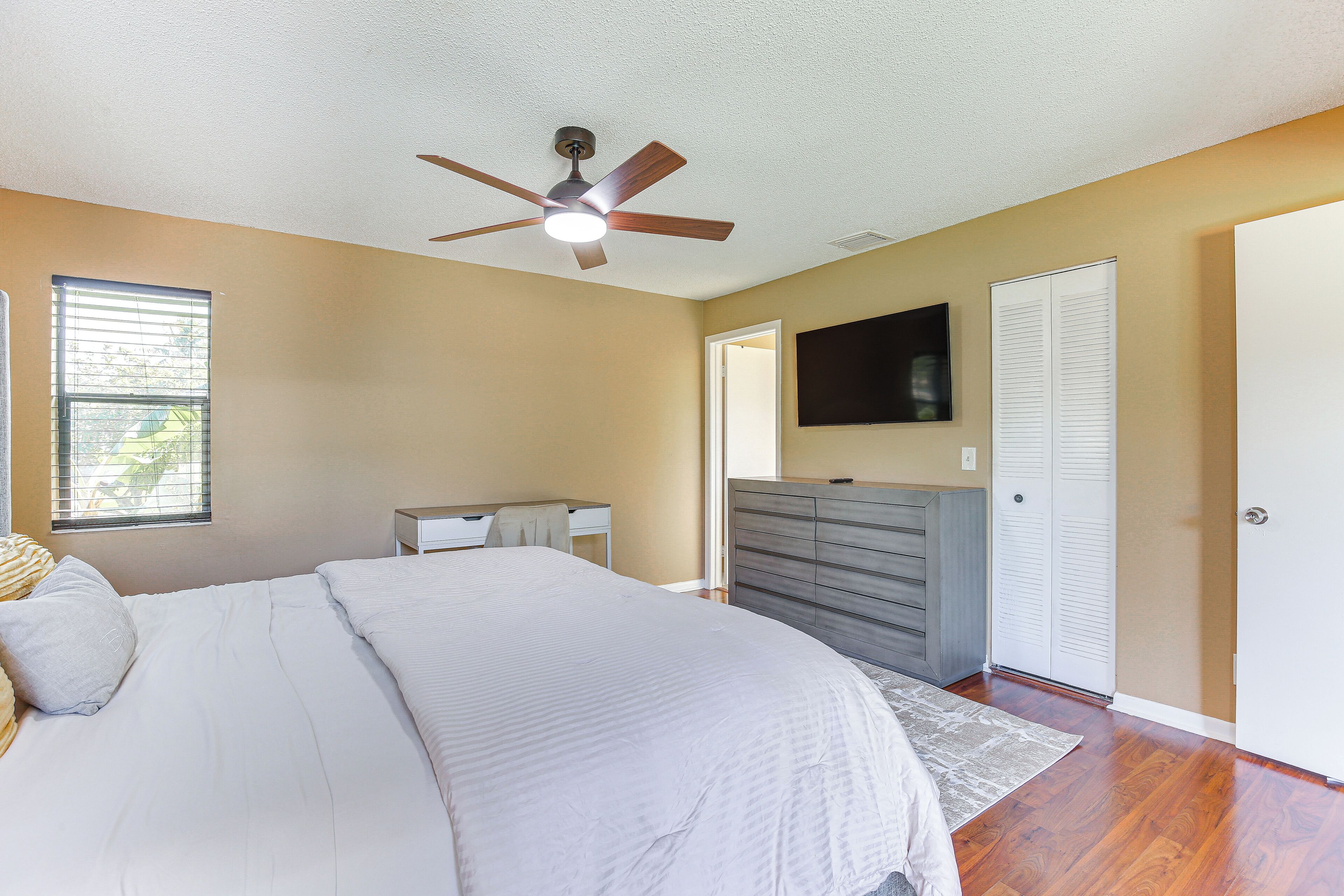 A bedroom with a bed, a desk, a dresser, a ceiling fan, and a TV on the wall.